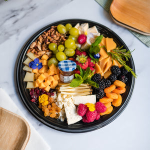 Assorted fruits, cheeses, and nuts on a black plate with a decorative background.