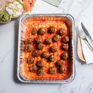 Aluminum pan with meatballs on a sweet chili sauce, placed on a marble surface with flowers and cutlery in the background.