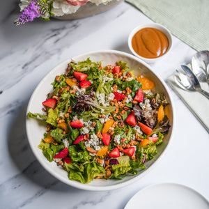 Colorful salad in a bowl on a marble surface with a side of dressing, utensils, and flowers.