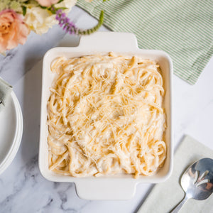 Baked pasta dish in a white dish on a marble surface with flowers and a spoon.