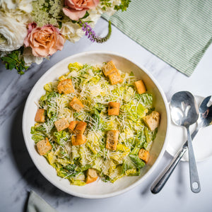 Salad with croutons in a white bowl on a marble surface with flowers and spoons.