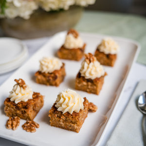 Small square desserts with whipped cream on a white plate, set against a blurred floral and tablecloth background.