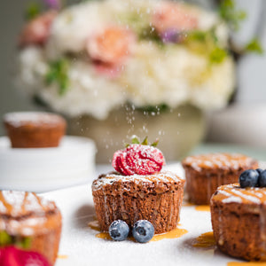 Brownies with berries on a decorative tablecloth, with a blurred floral arrangement in the background.