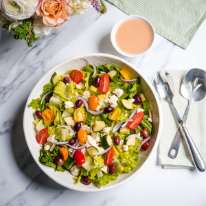 Colorful salad in a bowl with a side of dressing, flowers, and cutlery on a marble surface.