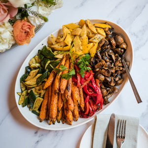 Assorted roasted vegetables on a plate with cutlery and flowers on a marble surface