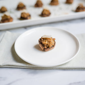 Stuffed mushroom on a white plate with a tray of more mushrooms in the background.