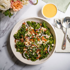 Kale salad with a side of dressing on a marble surface with flowers and cutlery.
