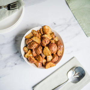 Bowl of roasted potatoes on a marble table with a floral arrangement and cutlery.