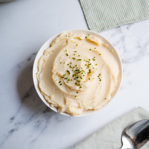 Bowl of mashed potatoes on a marble surface with flowers and a spoon.