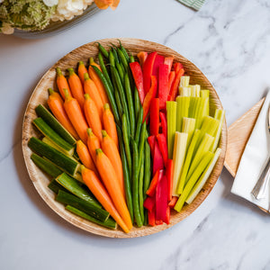 Crudité platter with long-stem baby carrots, haricots verts, red bell pepper strips, cucumber spears, and celery, arranged on a round wooden serving tray next to a floral centerpiece